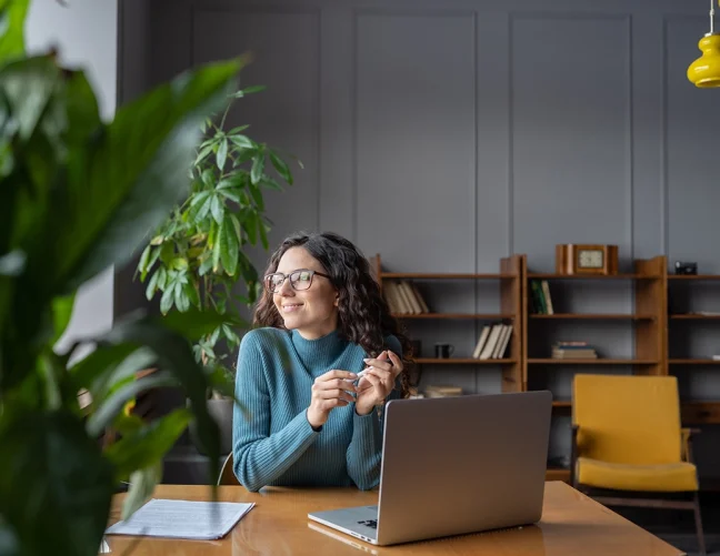 Woman with spectacles sat at a table with an open laptop, gazing out of the window.